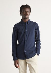 Marineblauwe button-up shirt met een gladde textuur en lange mouwen, voorzien van een klein logo op de borst en contrasterende knopen.
