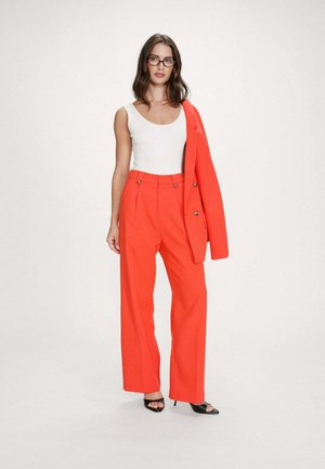 Femme portant des lunettes, un haut blanc sans manches, un pantalon large taille haute d'un orange vif, et un blazer orange assorti posé sur l'épaule.