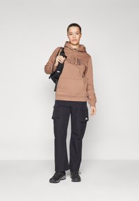 Felpa marrone con logo, pantaloni cargo neri con tasche e stivali neri con lacci. Il modello tiene una piccola borsa nera. Design e colori semplici.