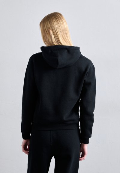 Blond inimene, seljaga seistes, mustas kapuutsiga särk ja mustades pükstes, heleda halli tausta ees.