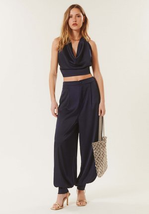 Femme portant un crop top sans manches bleu marine à col bénitier, un pantalon bleu marine ample avec des bas de jambes retroussés, tenant un sac à perles et des sandales dorées à talons hauts.