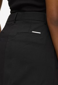 Čierne nohavice s presne strihom, hladkou textúrou, priliehavým pásom a malým strieborným akcentom na zadnej vrecku. Minimalistický dizajn.