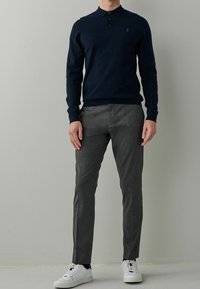 Dunkelblaues Langarm-Polohemd mit Kragen, drei Knöpfen und kleinem Logo, kombiniert mit grauen maßgeschneiderten Hosen und weißen Sneakers.