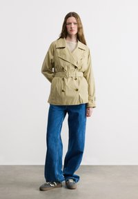 Jeune femme portant un trench-coat beige ceinturé, un jean bleu et des baskets grises, debout devant un fond blanc, les mains derrière le dos.