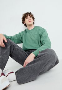 Groene katoenen sweatshirt met een ronde hals en geborduurd logo, gecombineerd met loszittende grijze denim broeken en witte sneakers met rode accenten.