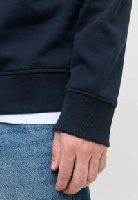 Granatowy sweatshirt z ściągaczami, charakteryzujący się gładką teksturą. Noszony na białej koszuli, zestawiony z jasnoniebieskimi dżinsami.