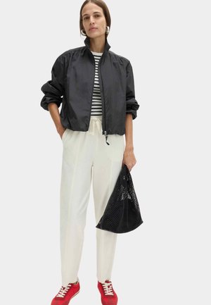 Donna che indossa una giacca nera con cerniera, camicia a righe, pantaloni bianchi, scarpe da ginnastica rosse, che tiene una borsa a rete nera su sfondo semplice.