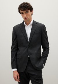 Traje de rayas en gris oscuro con solapa con muesca, cierre de un solo botón y dos bolsillos frontales, combinado con una camisa de vestir blanca. Textura suave.