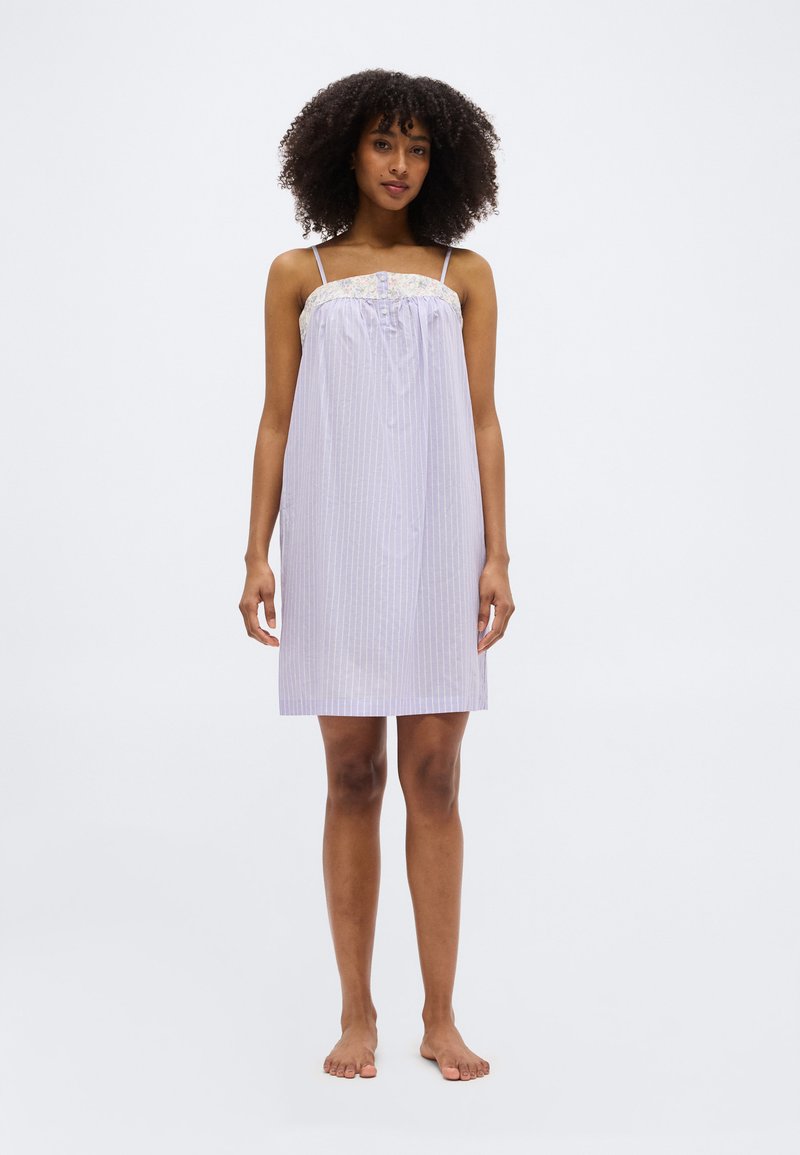 Γυναίκα με σγουρά μαλλιά που φοράει ένα μωβ φόρεμα χωρίς μανίκια, μήκους μέχρι το γόνατο, με λεπτές ρίγες και διακοσμητικά λουλούδια, ξυπόλυτη, σε λευκό φόντο.