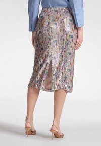 Kvinde iført en skinnende, flerfarvet paillet midinederdel med en slids bagpå, blå langærmet skjorte og beige høje hæle.