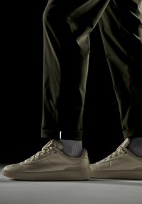 Beżowe sneakersy o gładkim, skórzanym wyglądzie, niskim profilu, zaokrąglonym nosku i teksturowanej podeszwie, zestawione z ciemnymi spodniami i jasnoszarymi skarpetkami.