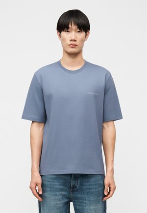 Bărbat tânăr cu păr negru și drept, purtând un tricou bleu cu mâneci scurte și un text mic alb pe piept și blugi albaștri.