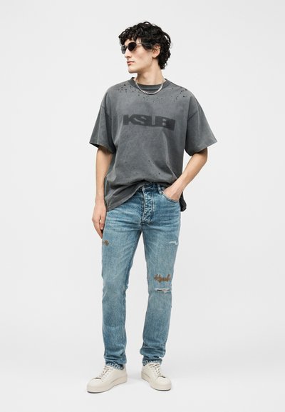 Νεαρός άνδρας με γυαλιά ηλίου, άνετο γκρι T-shirt Ksubi, φθαρμένα μπλε τζιν και λευκά αθλητικά παπούτσια, στέκεται με το ένα χέρι στην τσέπη.