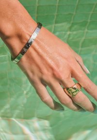 Sølvtekstureret armbånd og en guld, uregelmæssigt formet ring båret på en hånd med våd hud, nedsænket i lys turkis vand.