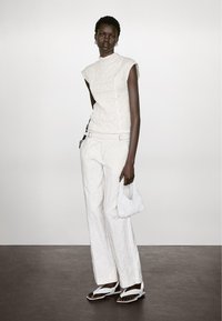 Witte textuurloze top zonder mouwen gecombineerd met losse witte broeken. Model houdt een kleine geweven witte tas vast en draagt witte platte sandalen met riempjes.