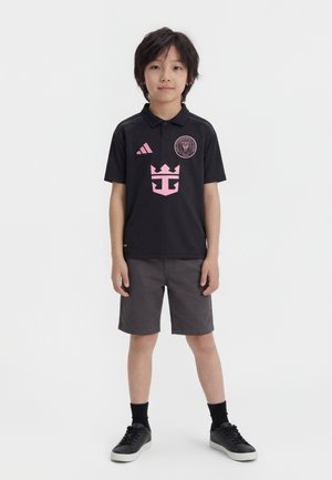 Noor poiss seisab näoga ettepoole, kandes musta polosärki roosade logodega, tumedad hallid lühikesed püksid, mustad sokid ja mustad tossud.