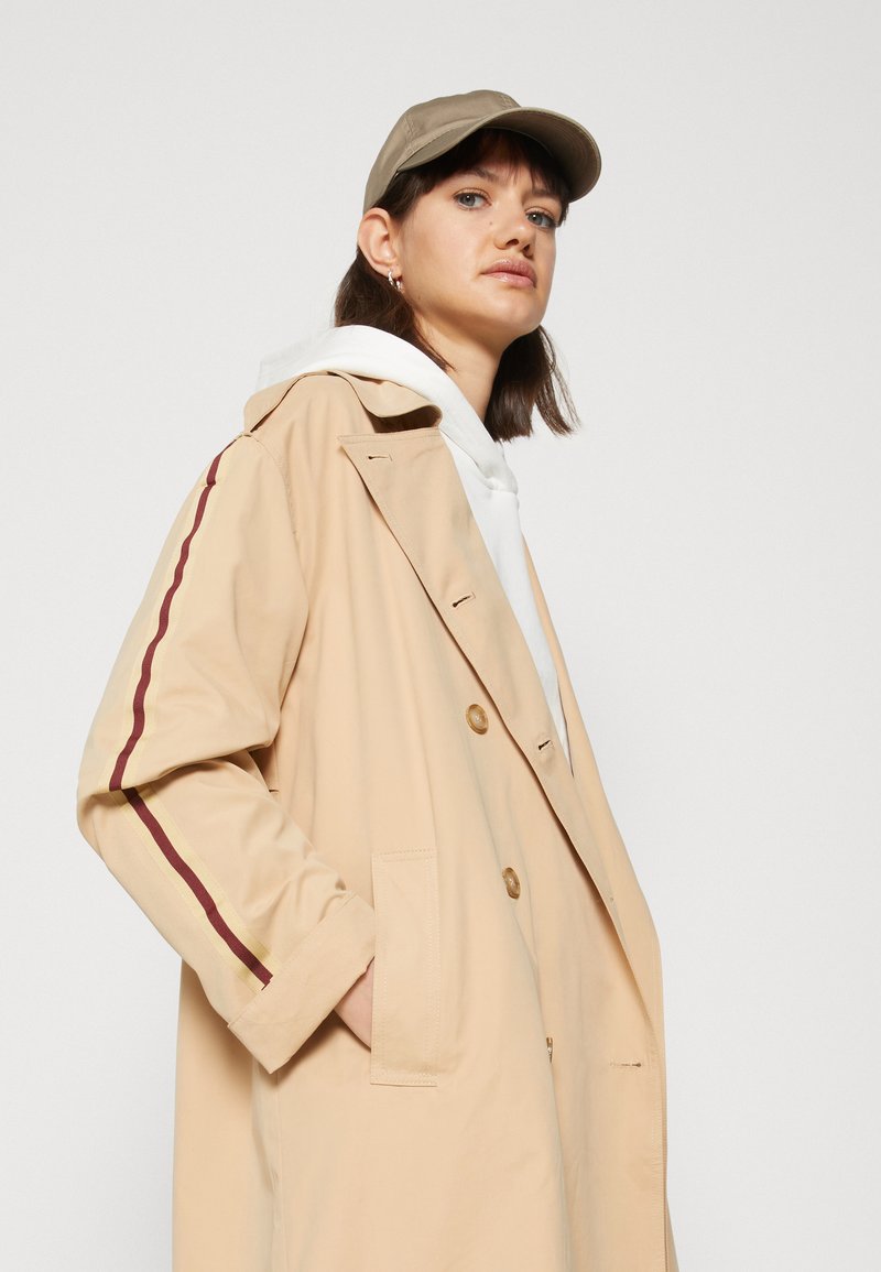 Mujer vestida con un abrigo trench beige con rayas rojas y amarillas en las mangas, sudadera blanca y gorra de béisbol beige mirando hacia adelante.