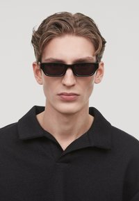 Occhiali da sole neri con lenti rettangolari e una spessa montatura nera, indossati da una persona in una camicia nera con colletto, che mostra un'espressione neutra.