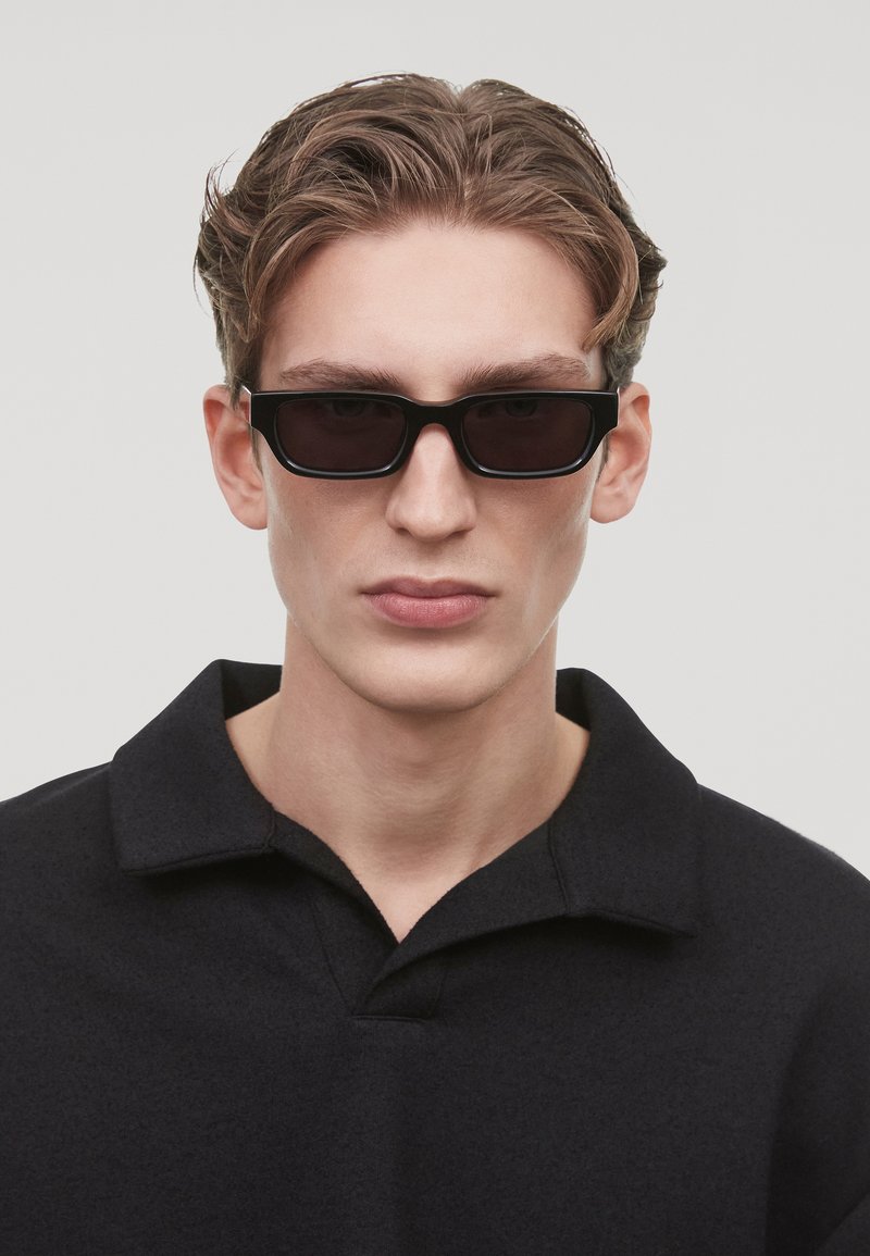 Occhiali da sole neri con lenti rettangolari e una spessa montatura nera, indossati da una persona in una camicia nera con colletto, che mostra un'espressione neutra.