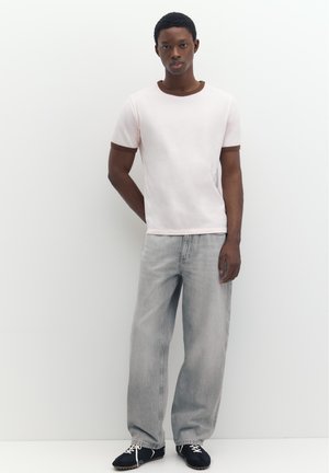Giovane uomo che indossa una t-shirt rosa pallido con rifiniture marroni, jeans larghi grigio chiaro e scarpe da ginnastica nere, in piedi contro un muro bianco.