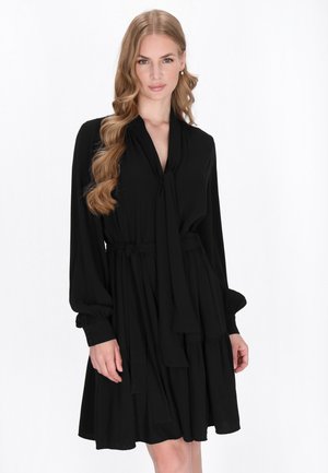 Robe noire faite d'un tissu doux, avec un col à nouer, des manches longues et un design de jupe à volants avec des poignets boutonnés.