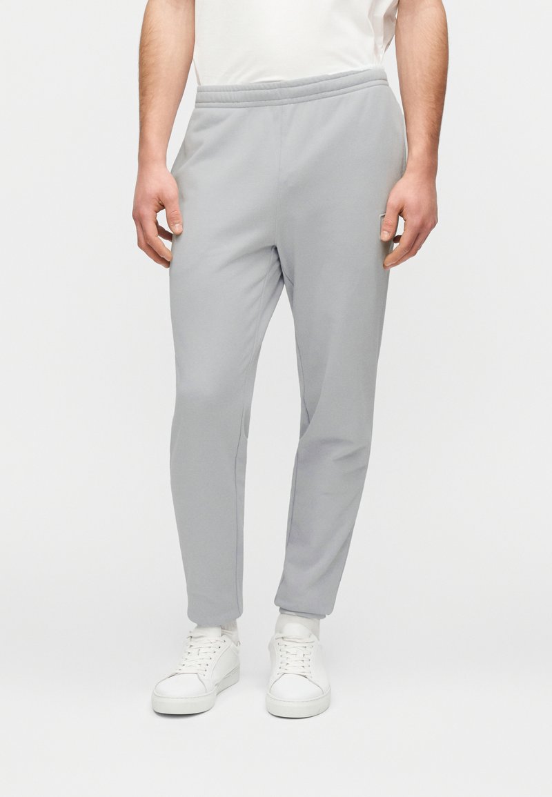 Ανοιχτό γκρι φόρμα με ελαστική μέση, σχεδιασμό στενών ποδιών και απαλό ύφασμα, σε συνδυασμό με λευκά αθλητικά παπούτσια.