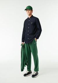 Camicia navy a bottoni, pantaloni sportivi verdi con rifiniture bianche, cappellino verde e sneaker nere. Il modello tiene una giacca verde abbinata.