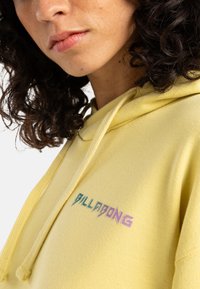 Yellow hoodie made of soft fabric, featuring a front logo in blue and pink gradient. Includes a drawstring hood and ribbed cuffs.
