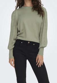 Vaaleanvihreä neulepusero, jossa on ribatty kaula-aukko ja koristeellinen reikäkuvio puffeilla hihoilla, yhdistettynä mustiin slim fit -farkkuihin.