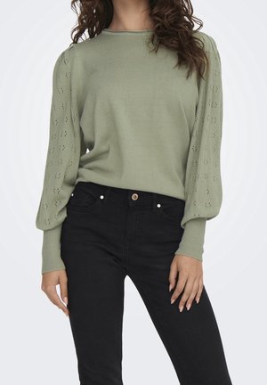 Vaaleanvihreä neulepusero, jossa on ribatty kaula-aukko ja koristeellinen reikäkuvio puffeilla hihoilla, yhdistettynä mustiin slim fit -farkkuihin.