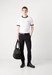 Bílý tričko s černým lemem, černé kalhoty a černé boty. Model drží černou látkovou tašku. Jednoduchý, čistý design bez vzorů.