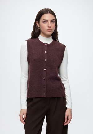 Femme aux longs cheveux foncés portant un gilet marron boutonné sur un haut blanc à manches longues et un pantalon marron foncé, debout devant un fond uni.