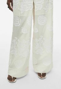 Pantalones de pierna ancha en tejido crema claro con prominente bordado floral blanco. Presentan un ajuste relajado y un acabado en el dobladillo a la altura de los tobillos.