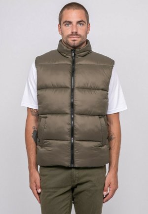 Homme aux cheveux courts et à la barbe, portant une veste matelassée vert olive sur une chemise blanche à manches courtes et un pantalon assorti vert olive, debout immobile.