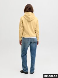 Persoon met krullend bruin haar, gekleed in een gele hoodie, blauwe jeans en zwarte schoenen, staand met de rug naar de kijker op een effen lichte achtergrond.