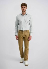 Homme se tenant debout, portant une chemise boutonnée gris clair, un pantalon en velours côtelé marron et des baskets blanches sur un fond blanc uni.