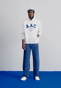 Pilkos spalvos hoodie su mėlynu tekstu, su raišteliu ir priekinėmis kišenėmis, derinama su plačiomis mėlynomis džinsinėmis kelnėmis ir baltais sportbačiais.