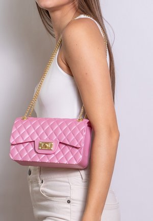 Vrouw draagt een wit tanktopje en broek, draagt een gewatteerde roze leren handtas met een gouden kettingriem over de schouder.