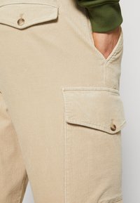 Beige kordfløyelsdungeri med en teksturert overflate, som har en brystlomme og sidelommer, dekorert med brune knapper.