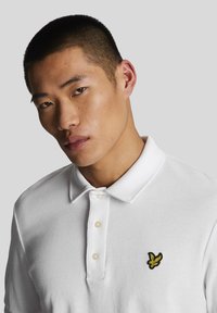 Polo bianco in tessuto strutturato, con due bottoni e un logo ricamato nero e giallo sul petto. Modello con capelli corti, sfondo neutro.