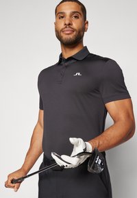 Polo nero con maniche corte, colletto a coste e logo bianco. Il modello tiene un bastone da golf, indossando un guanto bianco, su uno sfondo neutro.