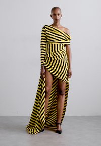 En axelklänning med gula och svarta ränder, figurnära liv, asymmetrisk fåll och sidoklyvning. Tillverkad av stretchigt tyg.