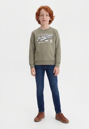Garçon roux debout, portant un sweat-shirt olive avec le texte « Jack & Jones », jean bleu et baskets marron sur fond blanc.