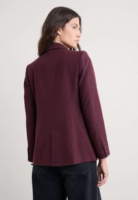 Blazer de lana burdeos con un corte ajustado, solapas con muesca y tres botones en el puño. La abertura trasera añade estructura al diseño.