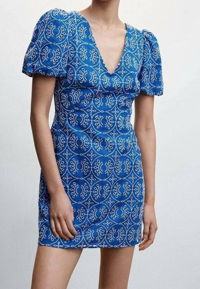 Femme portant une robe courte bleue avec des manches bouffantes et des motifs floraux circulaires blancs, debout devant un fond clair uni.