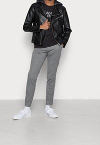 Giacca motociclista nera in pelle con cerniere argento, pantaloni grigi slim-fit e sneaker bianche. La giacca presenta una fodera interna con un motivo.