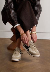 Beige sneakers met zwarte accenten, gemaakt van suede en mesh materiaal, vetersluiting, rubberen zool, gedragen met een bruine rok, houten sculptuur op de achtergrond.