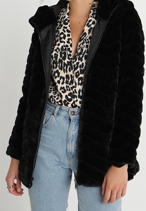 Vrouw draagt lichtblauwe spijkerbroek, een zwarte nepbont jas met rits en een witte blouse met luipaardprint en een diepe V-hals.