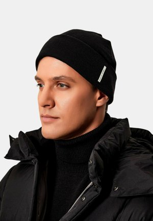 Berretto lavorato a maglia nero con risvolto, indossato insieme a un dolcevita nero e a un giubbotto imbottito, caratterizzato da bottoni a scatto e una finitura texturizzata.
