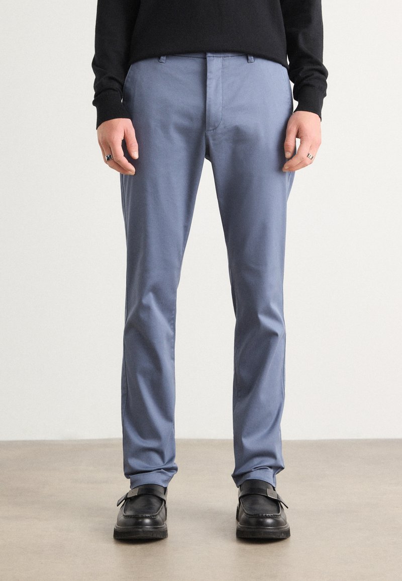 Pantalones ajustados de color azul claro, hechos de una tela suave. Presentan un corte recto y un ligero brillo. Combinados con zapatos de vestir negros.